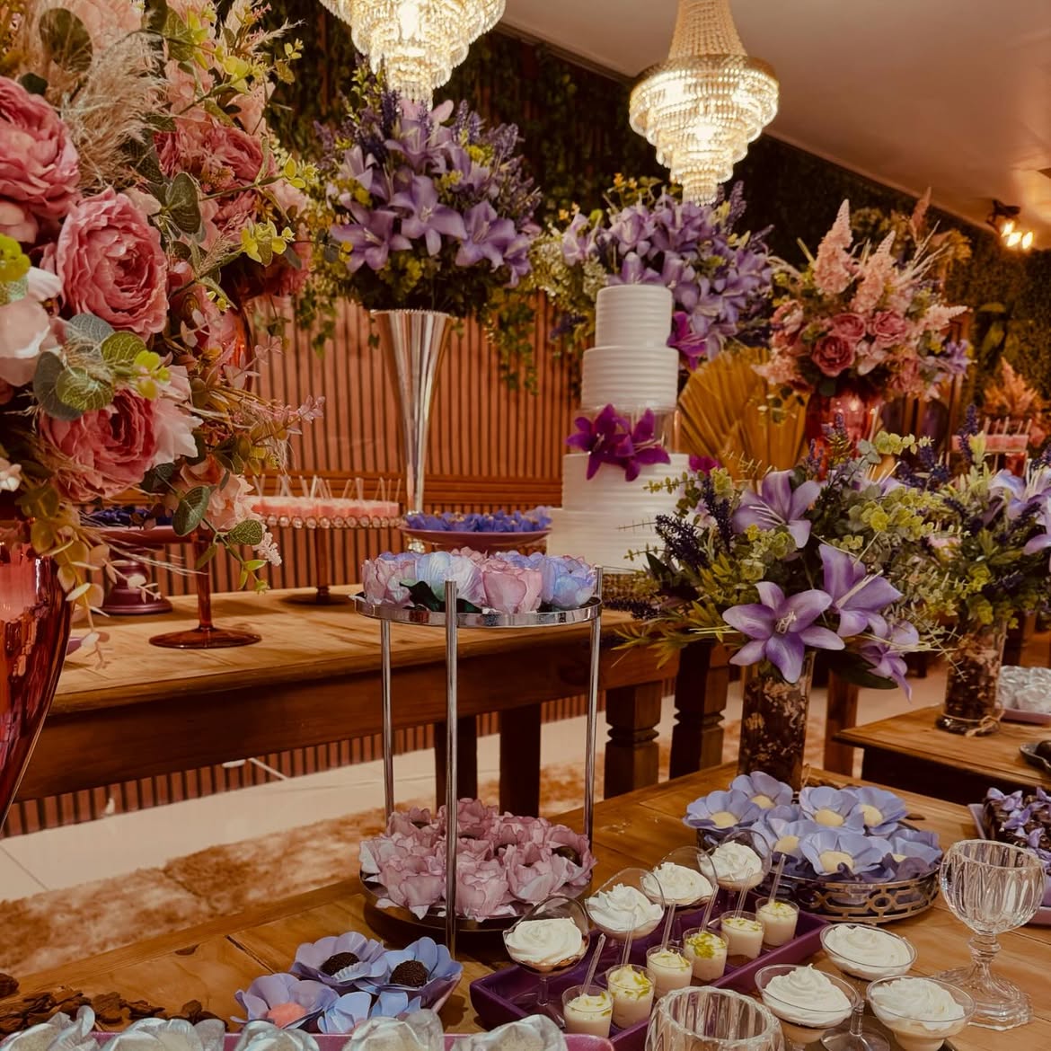 Mesa de doces e bolo temático com arranjos florais em tons lilás e rosa e lustres de cristal