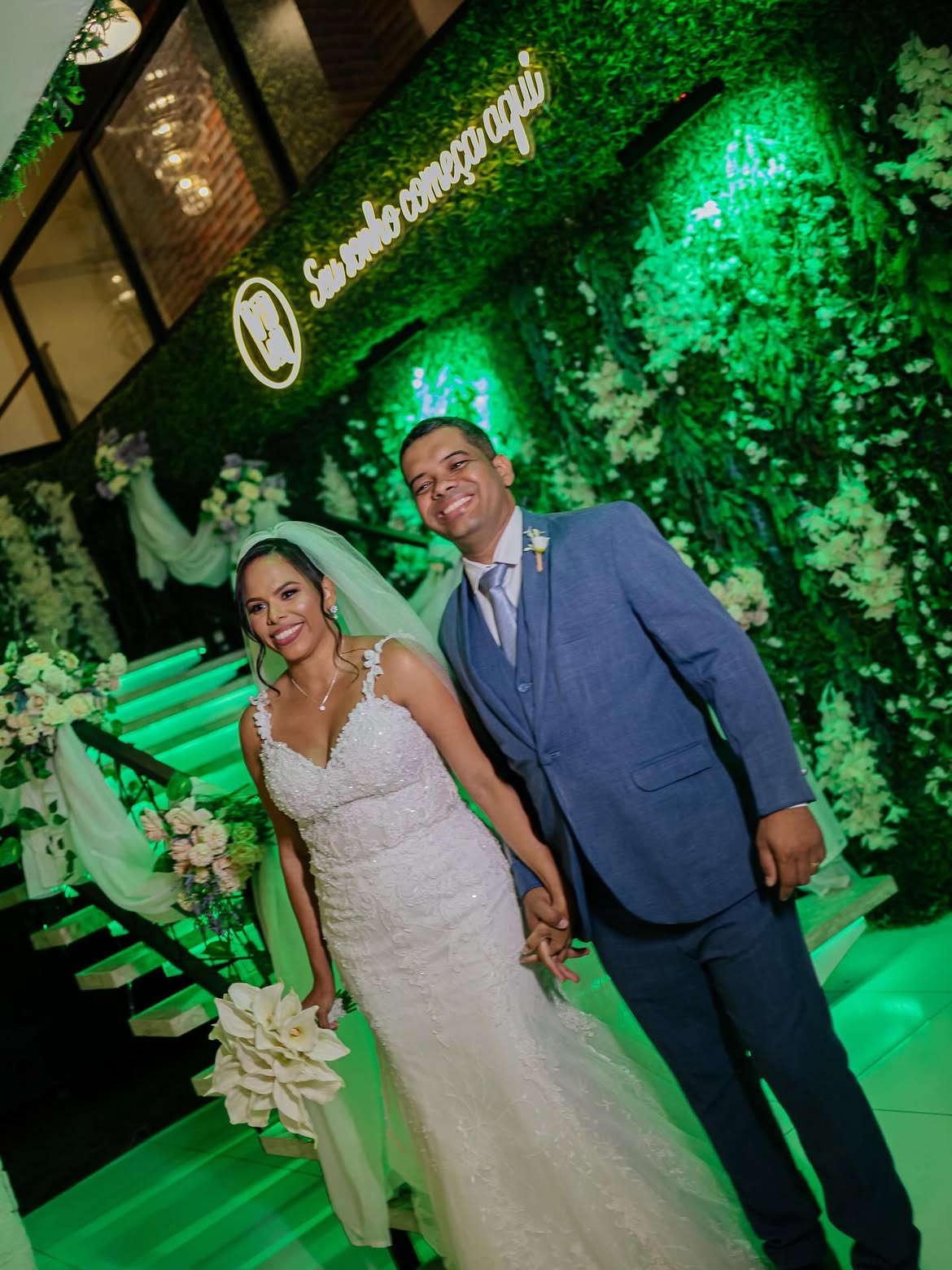 Noivos Nayara e Carlos na escadaria decorada com flores e letreiro em neon 'Seu sonho começa aqui'