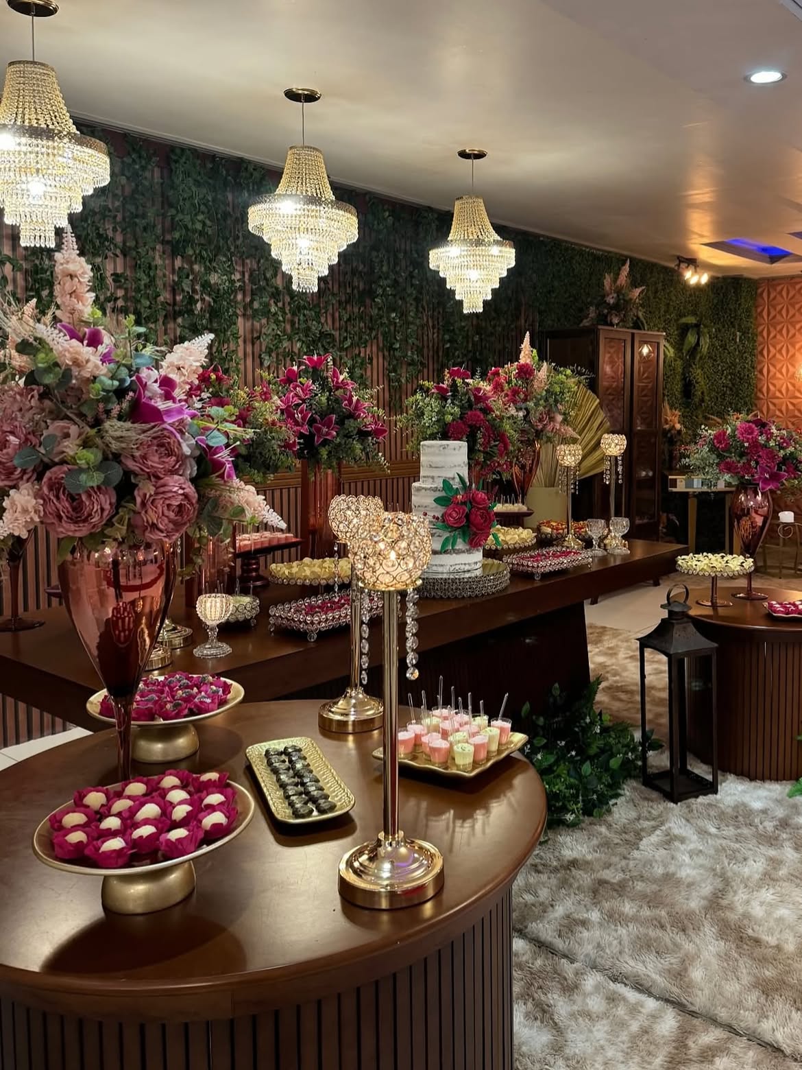 Mesa de bolo e doces finos com arranjos em tons de vinho, múltiplos lustres de cristal e painel ripado